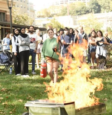 Person practicing use of a fire extinguisher on a real fire. Group of students watch.